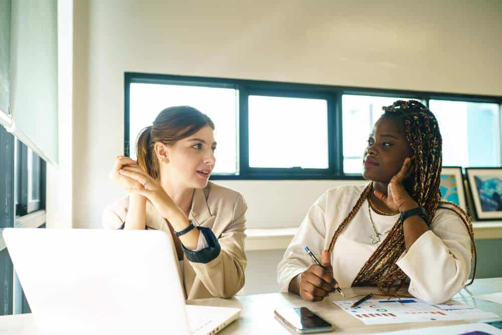 Two women discussing work with computer and paper in front of them.