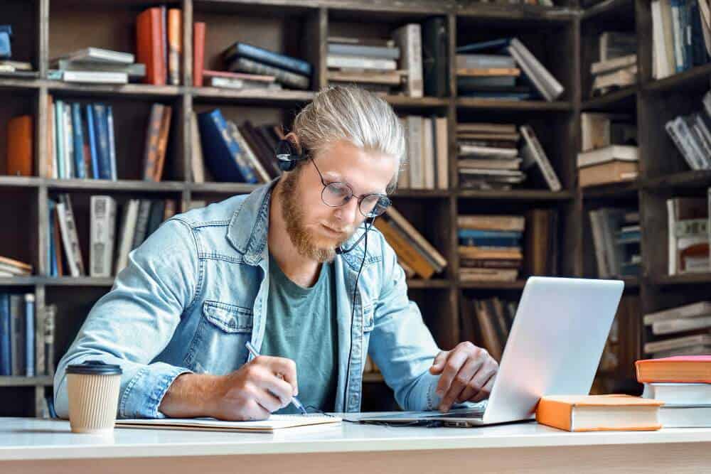 Man studies in library with laptop, headset and notebook.