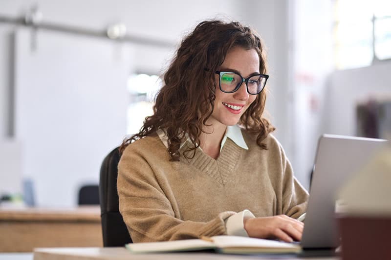 A professional woman with curly hair, wearing glasses and a beige sweater, smiles while working on a laptop in a modern office setting. This image represents the process of selling an online course, highlighting the importance of strategy and marketing.