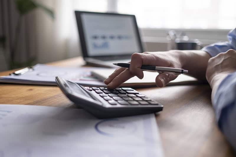 A close-up of a person using a calculator while holding a pen, with financial documents and a laptop in the background. This image represents the process of determining the best pricing strategy for selling an online course