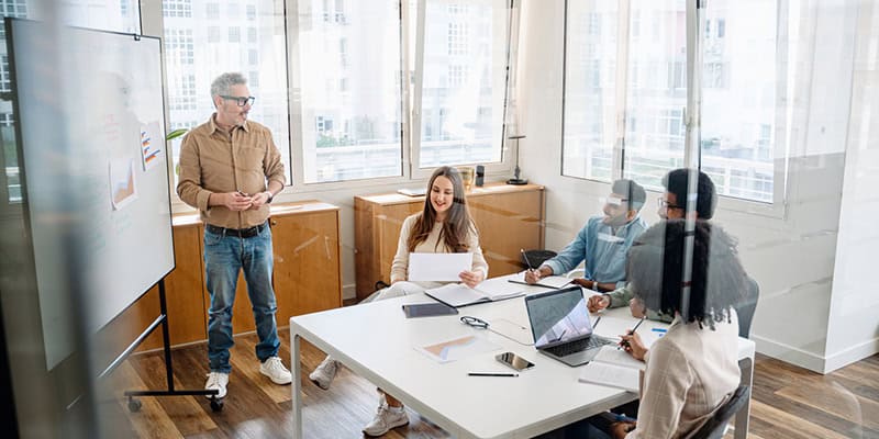 A diverse team of professionals collaborating during a strategy meeting, with a trainer leading the session in a modern office setting. The focus is on teamwork, learning, and development.