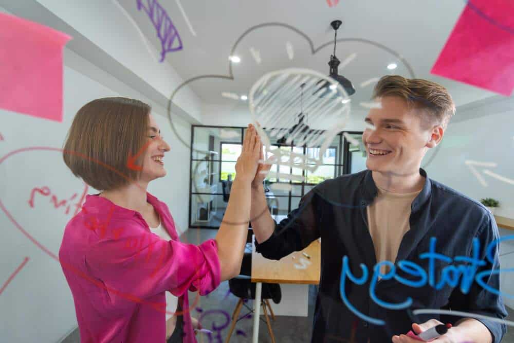 Woman and man colleagues giving high five in front of clear board.