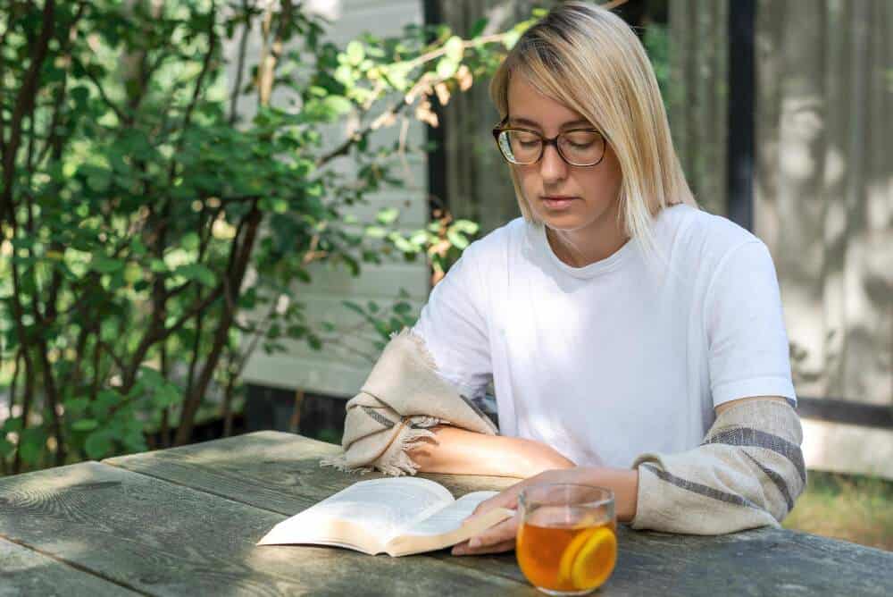 A young woman sitting by an outdoor table while reading and drinking lemonade