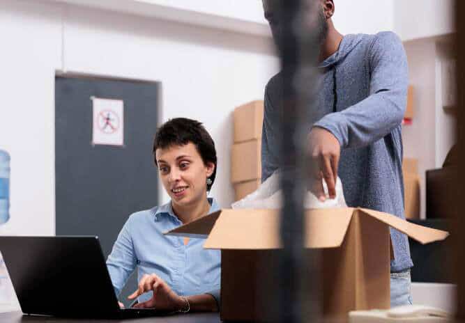 Person sitting on a desk with a computer while another person in unwrapping a box