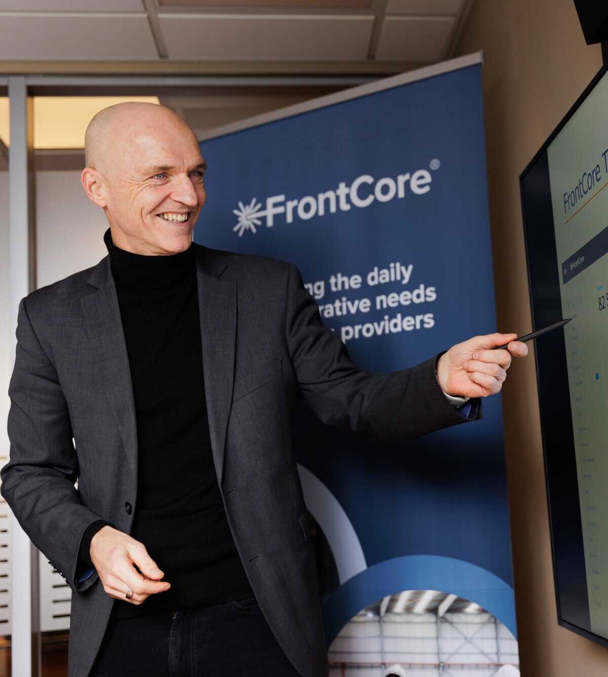 Smiling person in a blazer pointing at a wall-mounted screen showing the FrontCore Training Management dashboard.