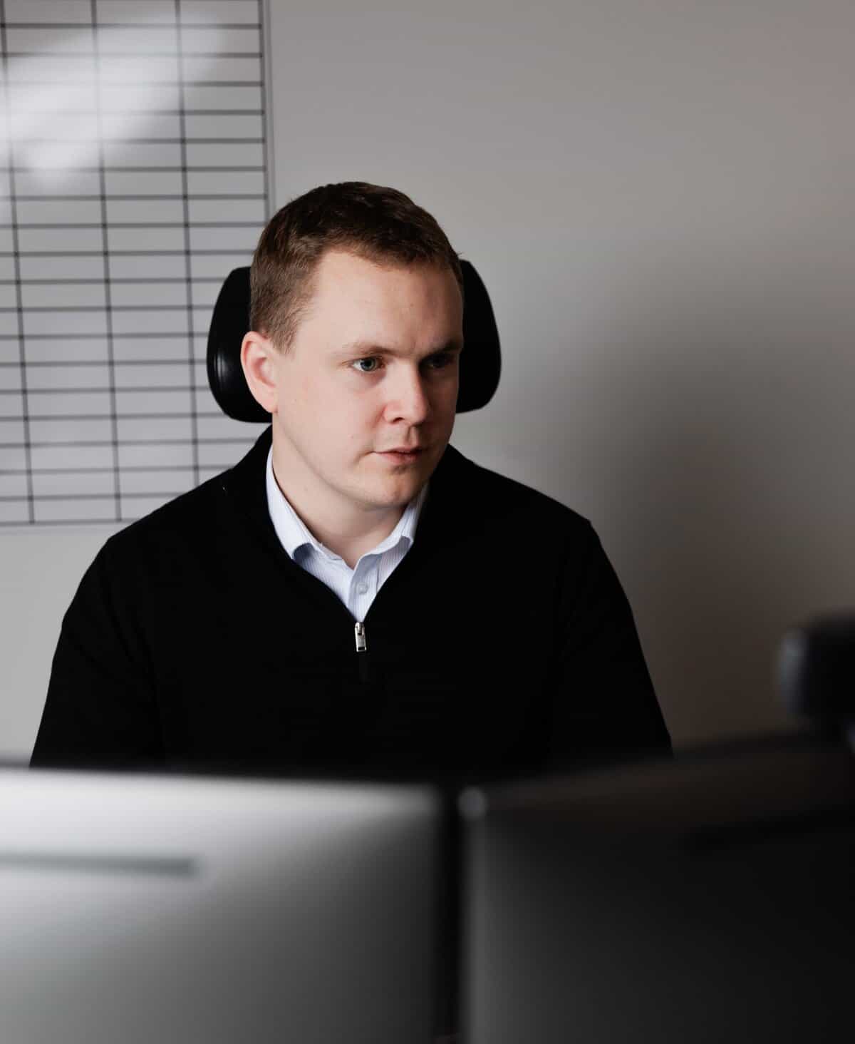 Person seated at a desk working on dual monitors, with planning grids on the wall in the background.