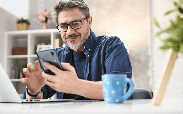 Man signing up for a construction course through his phone Smiling adult uses his smartphone next to a laptop and mug to register for a construction training course online.