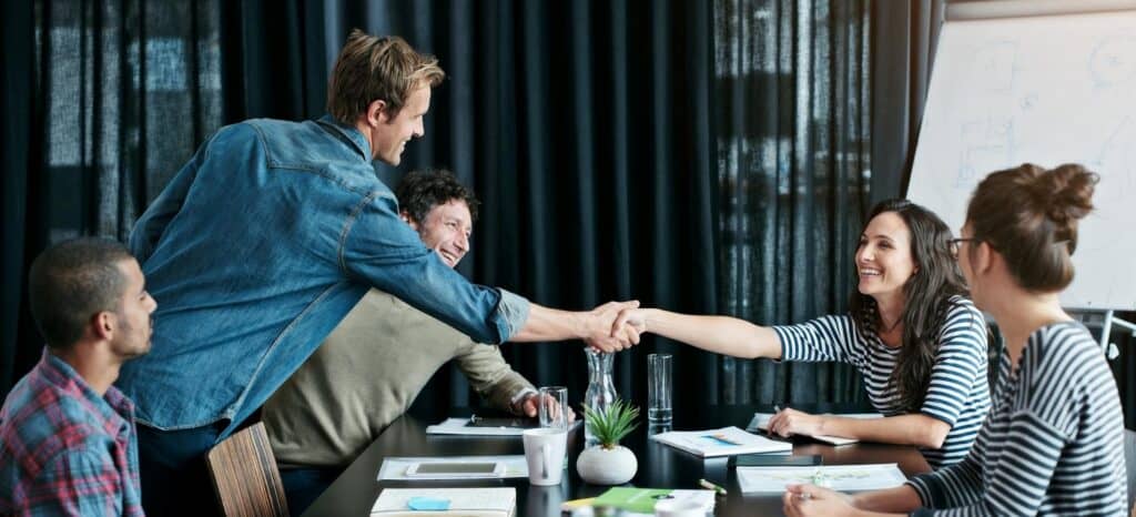 Four people are sitting around a table in an office, with a man handshaking one of the women.